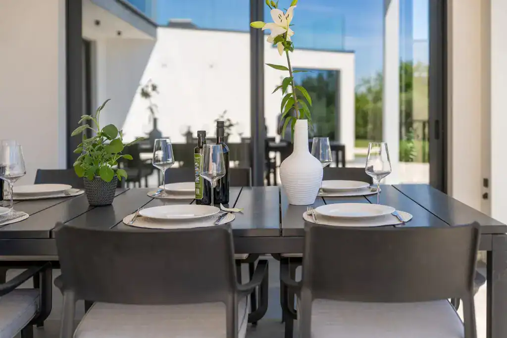 Überdachte Terrasse der Villa Nura mit gedecktem Esstisch, Lilie und Glasfront