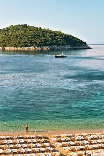 Strand mit Liegestühlen, zwei Personen im Wasser und Boot vor bewaldeter Landspitze